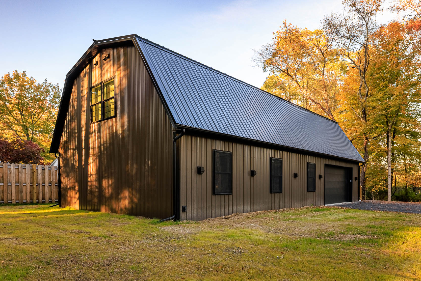 Rear angle of brown gambrel garage with black roof and warm autumn trees